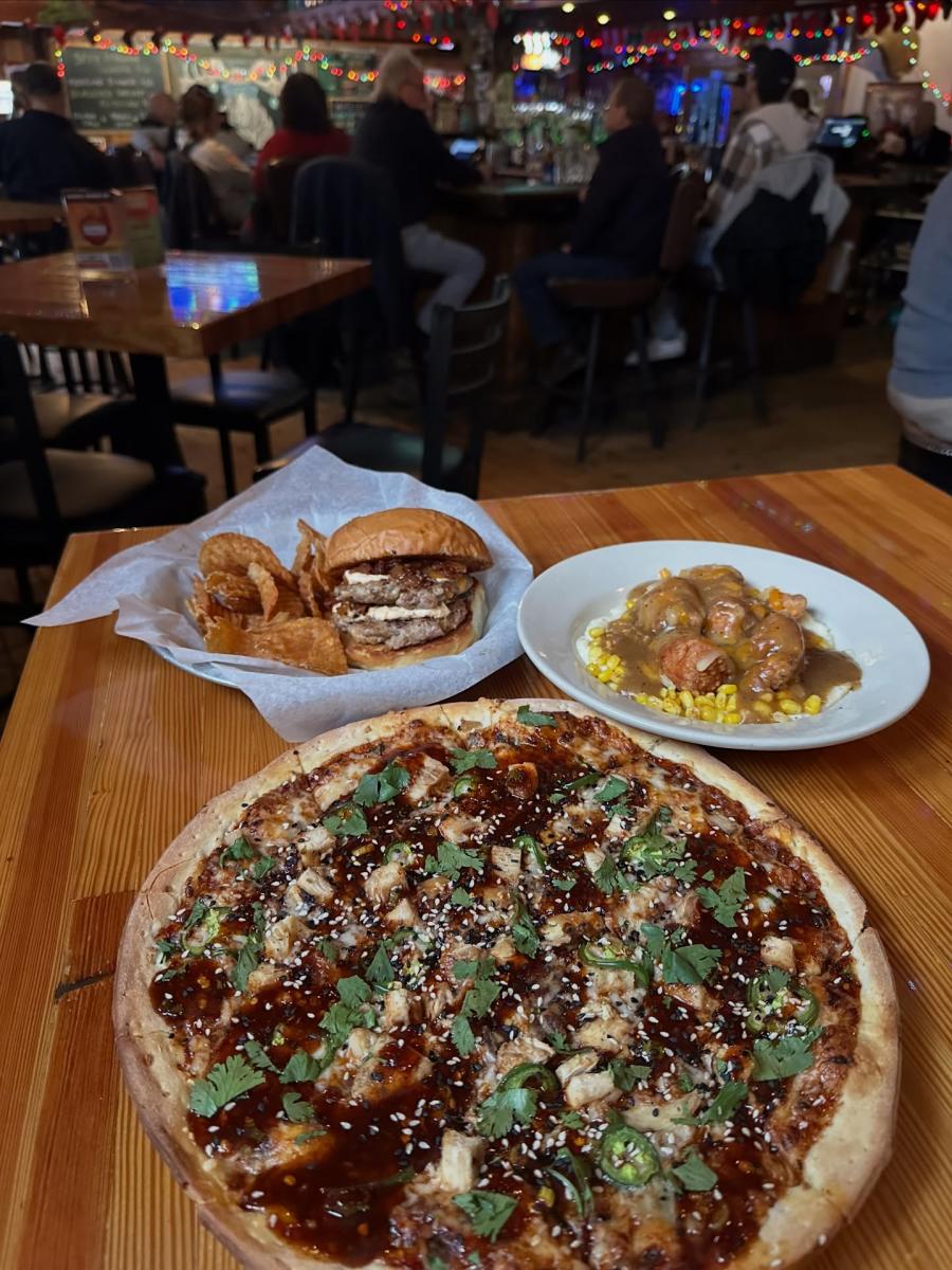 A pizza, a burger with house made chips, and a bowl of meatballs sits on a wooden table at Louie's Trophy House.