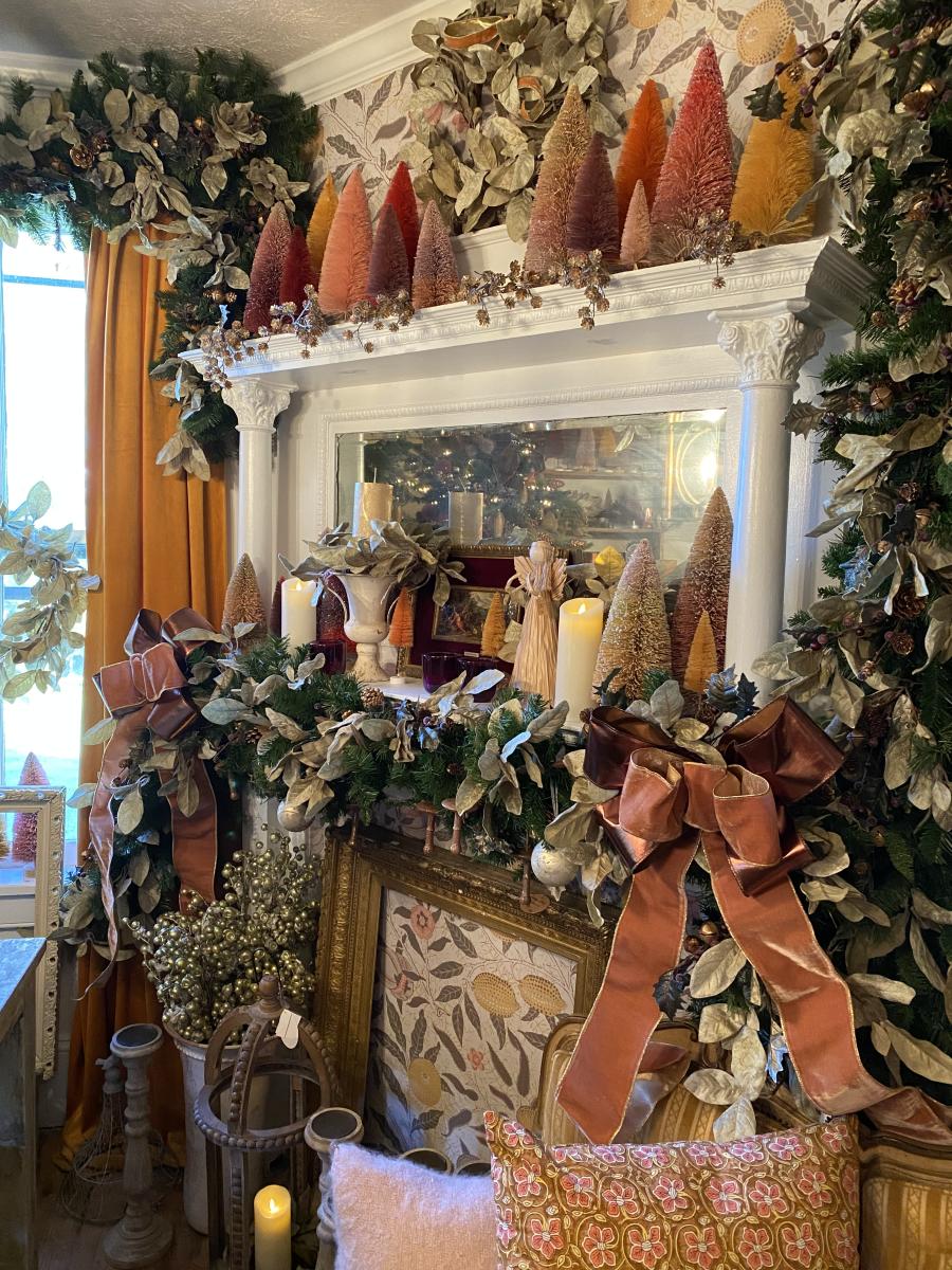 A white fireplace mantle is decorated with small colorful Christmas trees and green and white garland for sale during the Lucketts Holiday Market