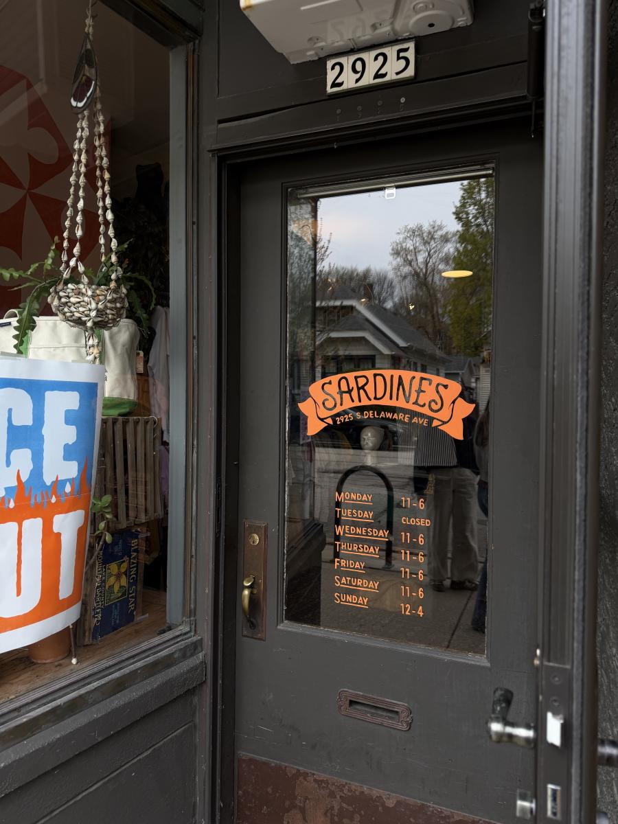Black storefront door for Sardines vintage shop with orange lettering listing hours, address “2925 S Delaware Ave,” and a weekly schedule, next to a display window with plants and retail items.