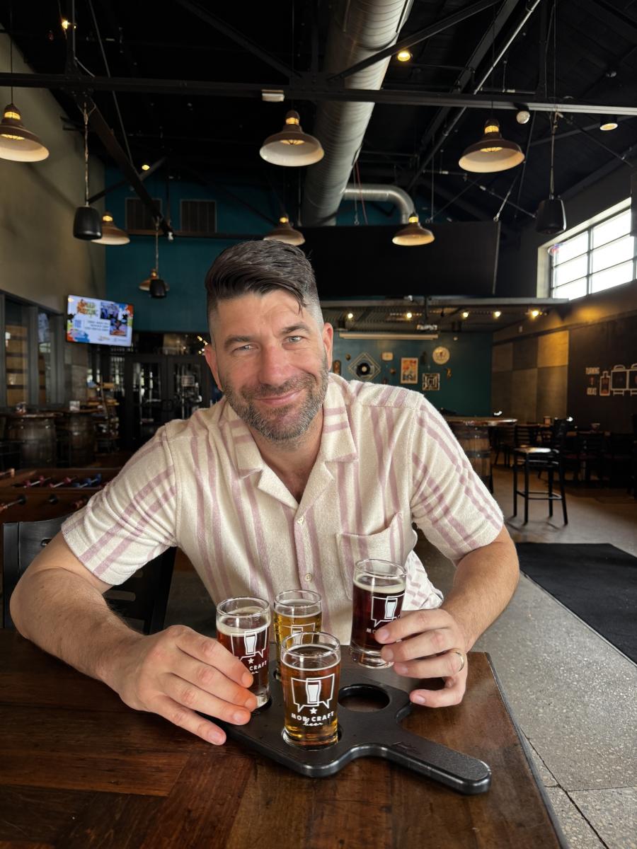 A smiling man holds a glass of beer while seated at a table with a flight of four craft beers at MobCraft Beer in Milwaukee.