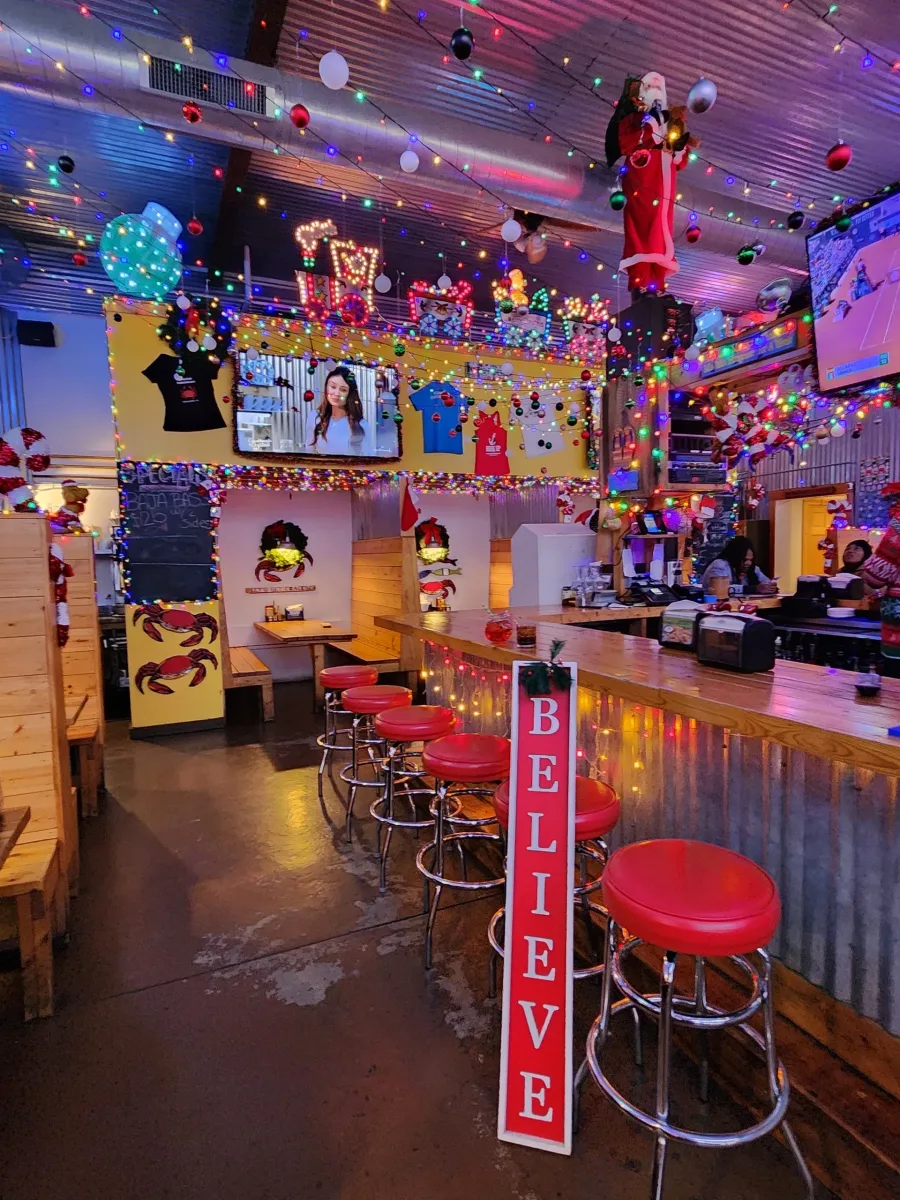 A brightly decorated bar interior filled with colorful holiday lights, ornaments, and themed displays. Red cushioned bar stools line a corrugated metal bar with twinkling lights hanging along the front. A tall sign reading Believe leans against the bar. Above the seating area are Christmas figures, strings of lights, and shirts displayed on a yellow wall. Two televisions are mounted near the ceiling while wooden booths sit along the left side of the room. The entire space feels festive and full of playful seasonal decor.