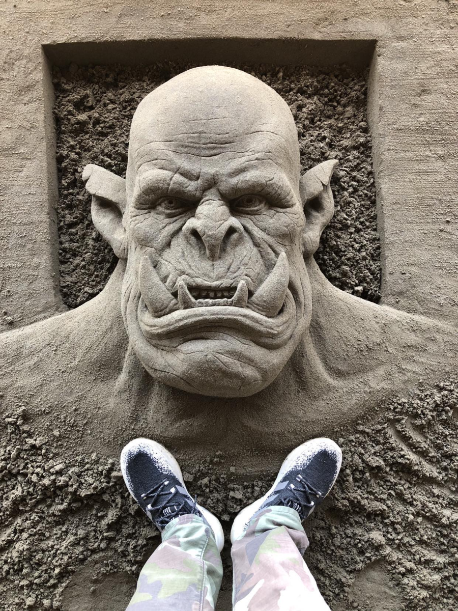 Sand sculpture in the ground showing a monster with large tusks' face. Below the ogre's neck are a set of feet standing over it taking the photo.