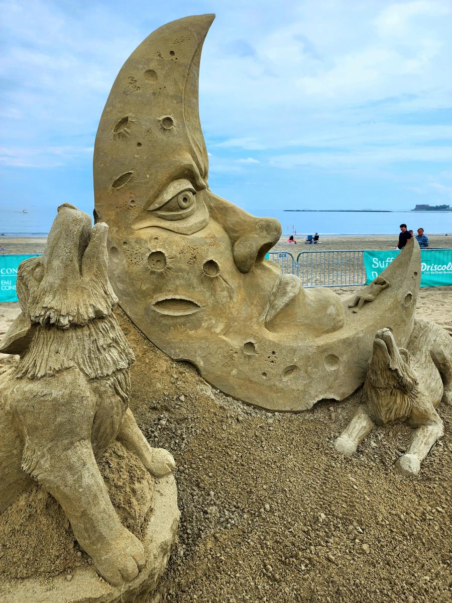 Sand sculpture depicting a crescent moon with a face surrounded by two howling wolves.