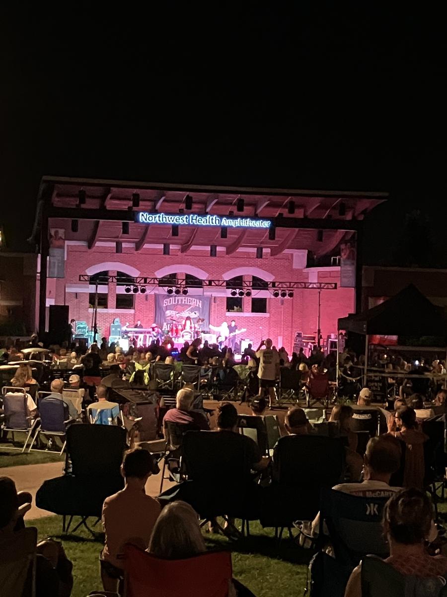 It is nighttime. A crowd of people sit in lawn chairs in front of a lit stage.