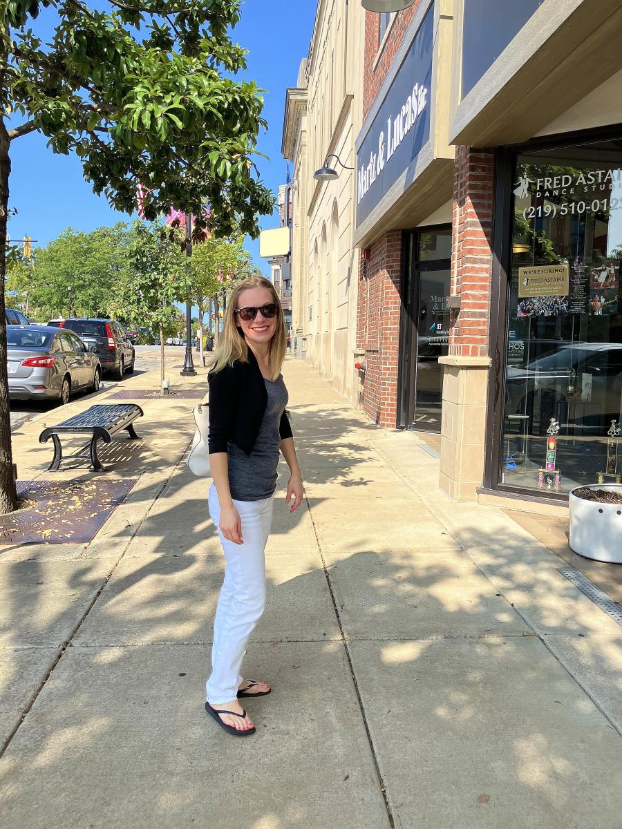 A woman with sunglasses stands on a sidewalk in front of storefronts.