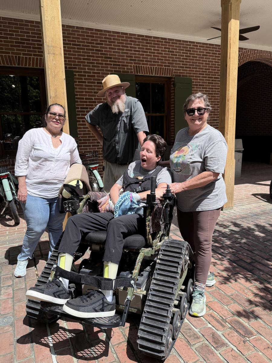 Christy and PJ Paulsell, joined by one of PJ's caregivers and Ranger Garrett, pose beside the all-terrain TrackChair at Fontainebleau State Park's Visitor Center.