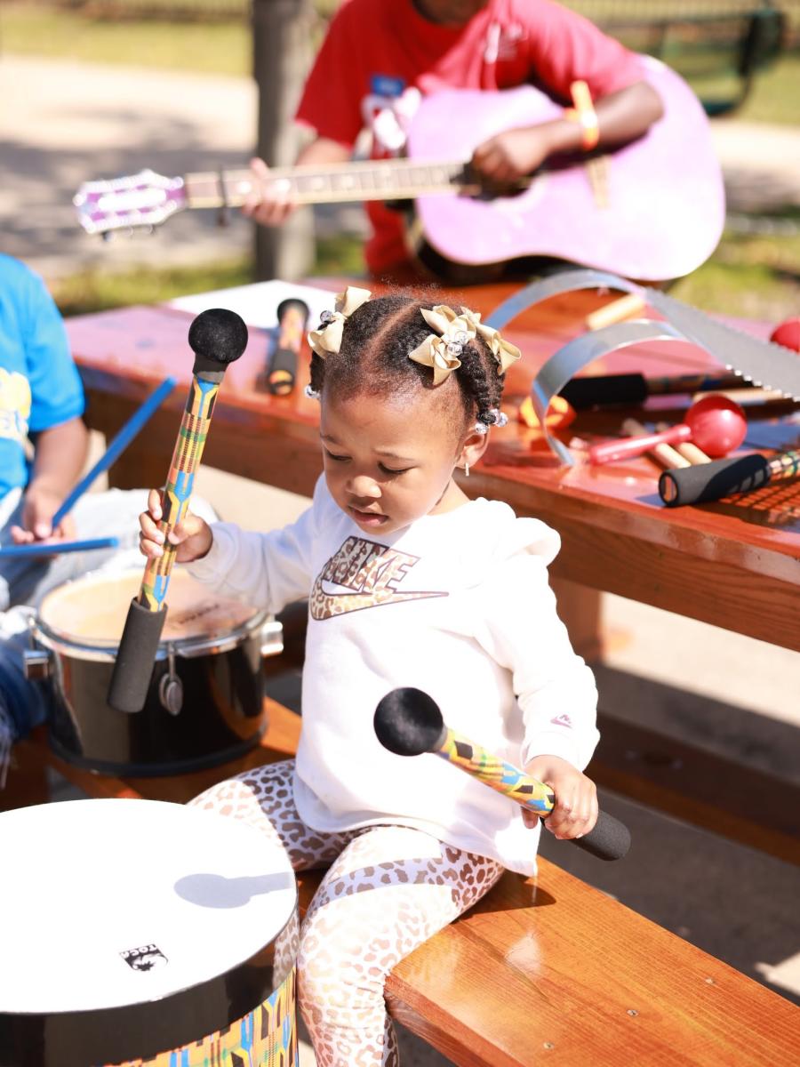 A young child plays a drum with colorful mallets during a hands-on music activity at NOLA Zydeco Fest in Slidell, with a guitar player and other instruments nearby.