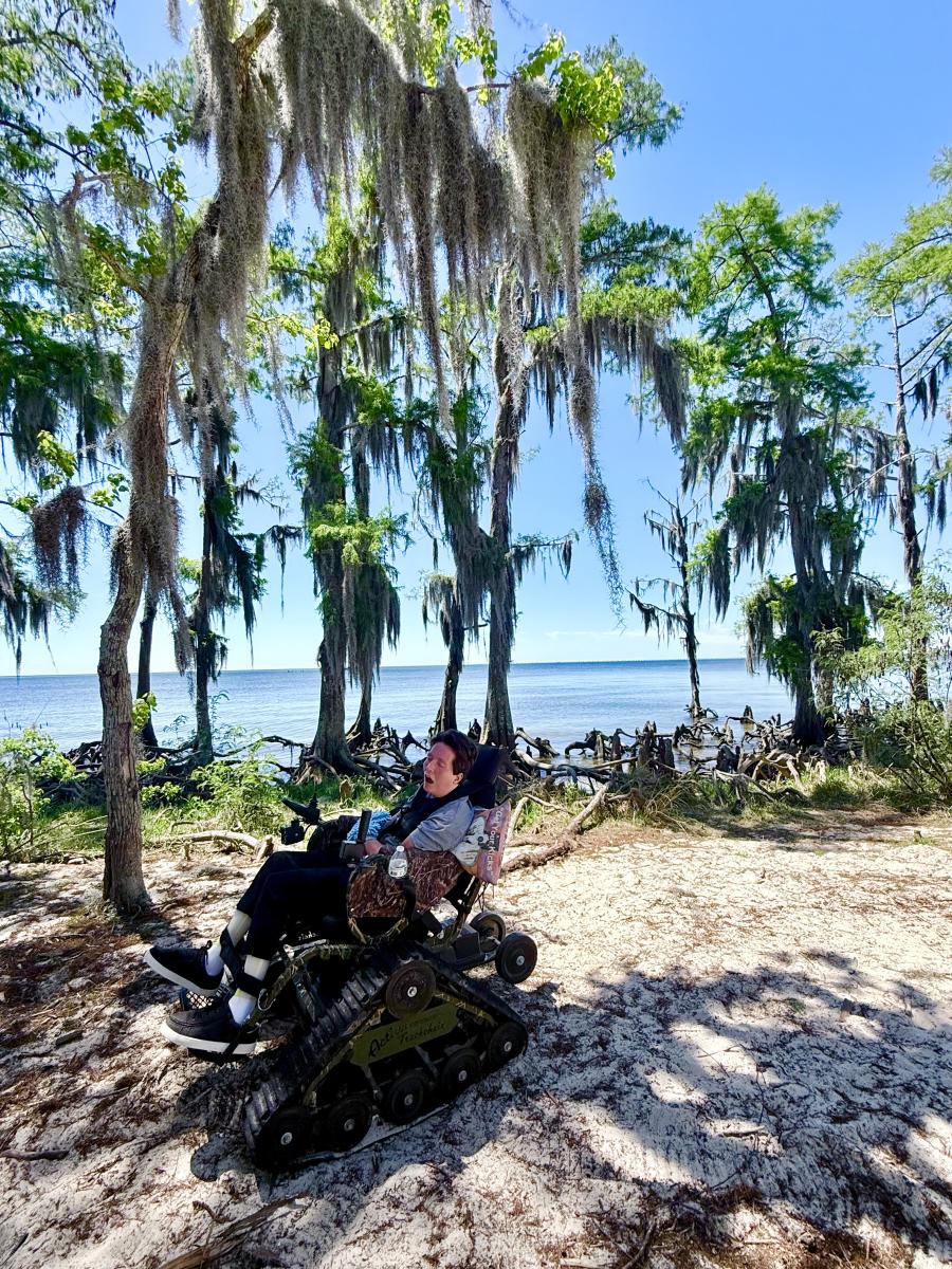 PJ Paulsell rides an all-terrain TrackChair along the sandy shoreline of Lake Pontchartrain at Fontainebleau State Park, demonstrating accessible outdoor recreation.