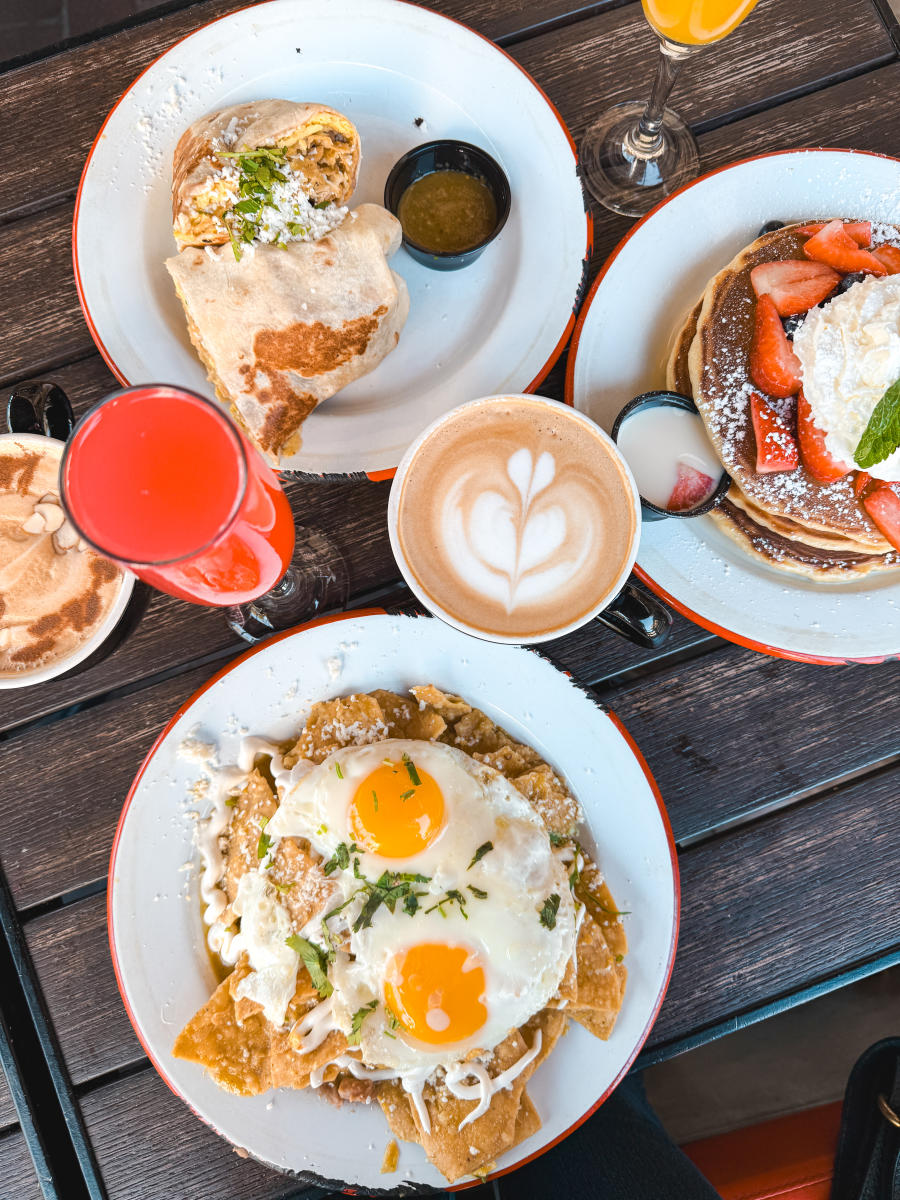 Overhead view of a brunch table at Cafe Cultura in Santa Ana featuring chilaquiles topped with eggs, strawberry pancakes with whipped cream, a breakfast burrito, lattes, and colorful brunch drinks.