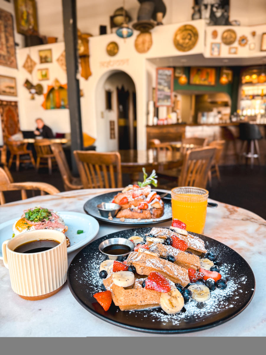 Brunch spread inside Artists Village Cafe in downtown Santa Ana featuring French toast topped with fresh berries, avocado toast, coffee, and orange juice in an eclectic café setting.