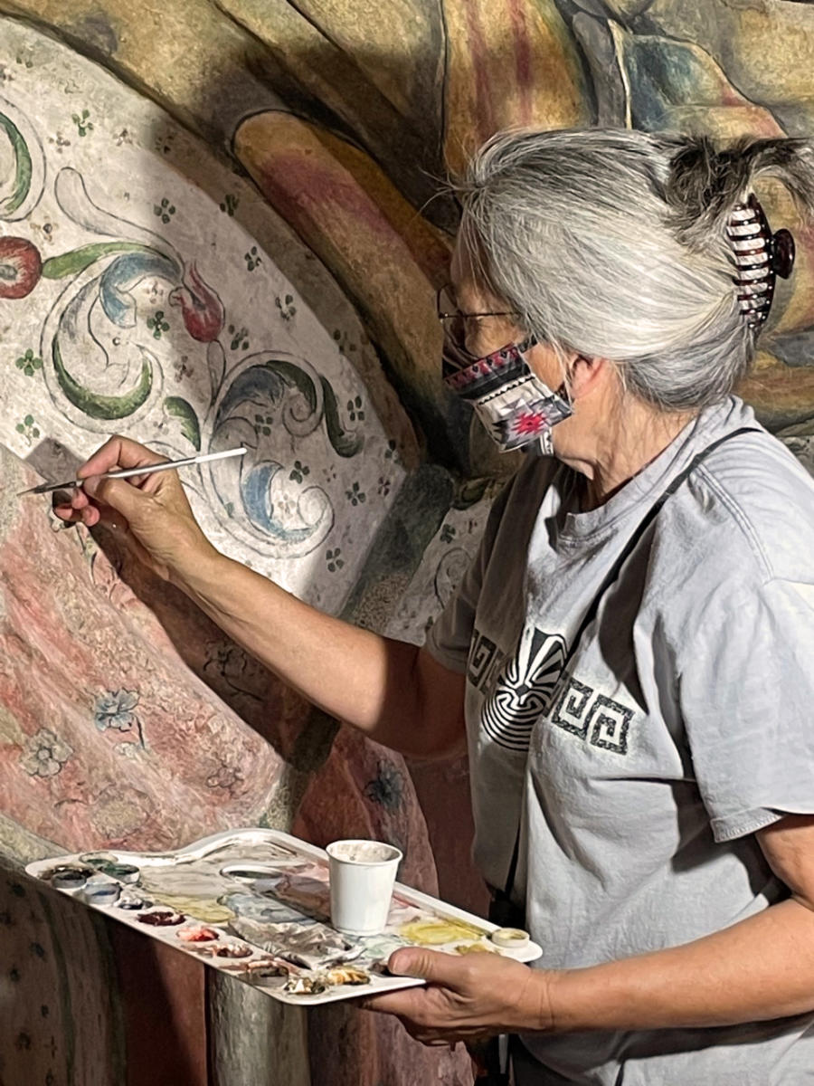 Woman holding an easel with paints and a small paintbrush restoring details on an old wall