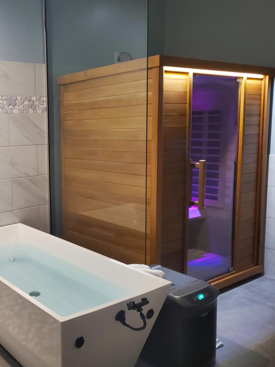 The camera overlooks a room with a shower (out of frame to the left); an angular bathtub full of water (perpendicular from the wall); and a wooden sauna with a glass door, lit from the inside with dim purple lights.
