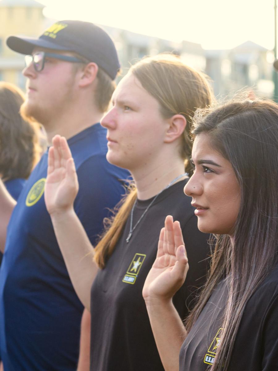 A group of young men and women are sworn in to the U.S. military. The camera focuses on a row of four initiates (three women, one man) raising their right hands to be sworn in.