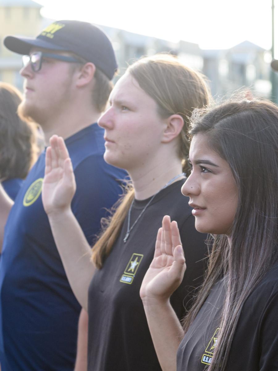 A group of young men and women are sworn in to the U.S. military. The camera focuses on a row of four initiates (three women, one man) raising their right hands to be sworn in.