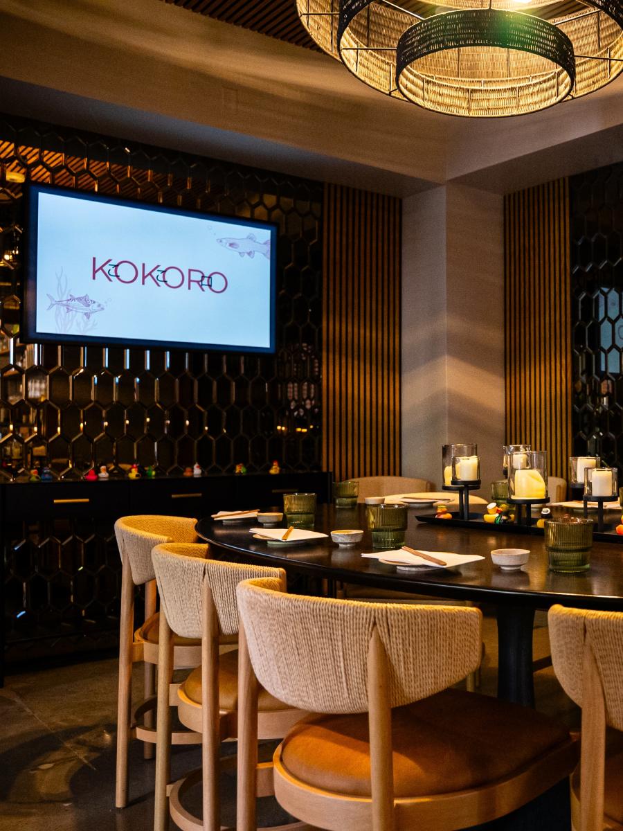 The camera overlooks a private dining room with an oval table made of dark wood, set with small water glasses, chopstick-laden plates, and glass-rimmed candles. Though empty, the room can seat eight. On the back left wall is a TV featuring the restaurant's name and logo.