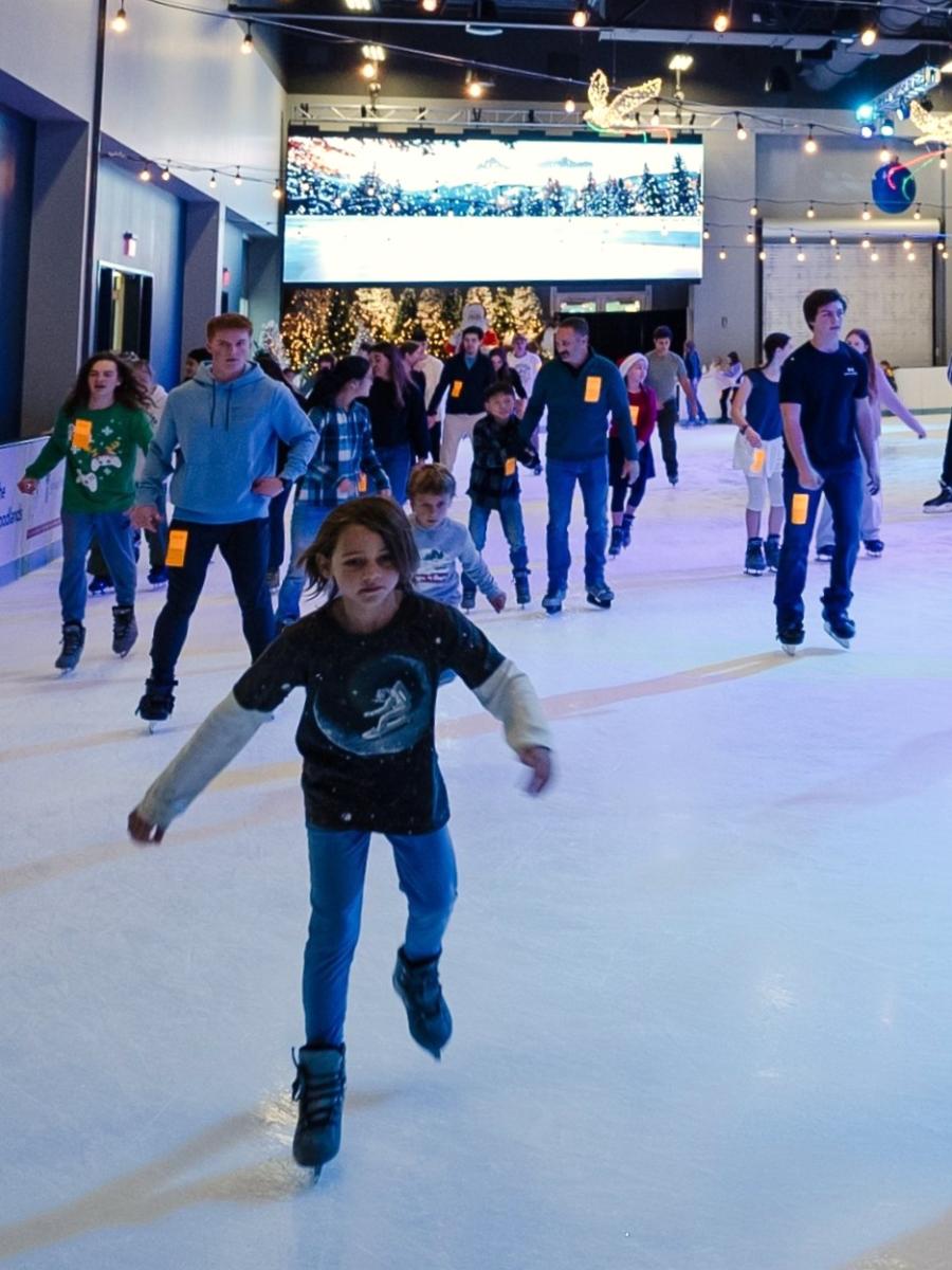 A boy (maybe 10 years old) skates toward the viewer at The Woodlands Ice Rink. Behind him are other kids, teens, and families skating around the rink. On the wall in the back is a large screen projecting a wintery nature scene.