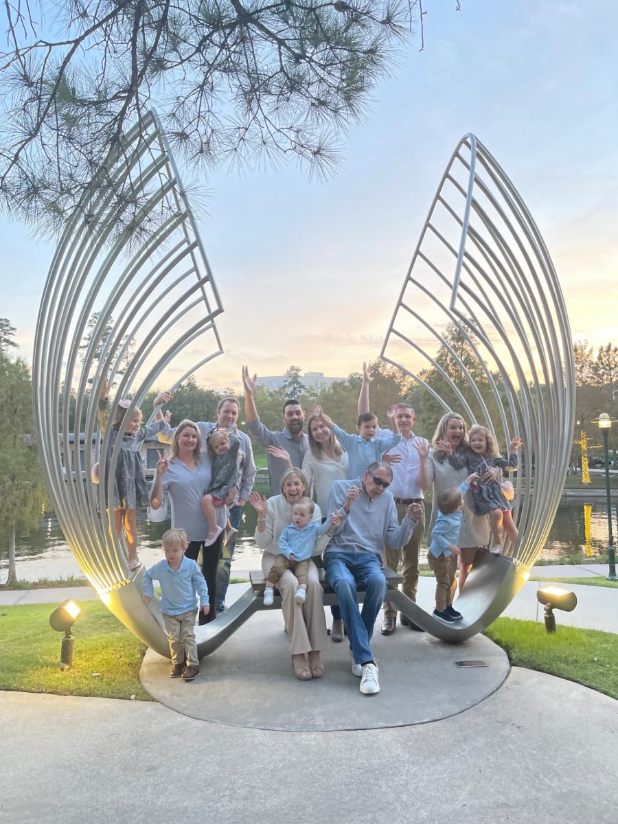 The camera faces the Memorial Hermman The Woodlands Medical Center's "Proud Souls" art bench, which looks like two large metal waves or wings spreading inward over the bench. Sitting on the bench is Linda Nelson and her husband. Behind and around them are their three children with their partners and seven children. Everyone is dressed in dressy-casual clothes in some shade of white, gray, or pale blue, and everyone (except a few runaway toddlers) is smiling and waving at the camera. In the background is The Woodlands Waterway and a beautiful wooded area on the opposite shore.