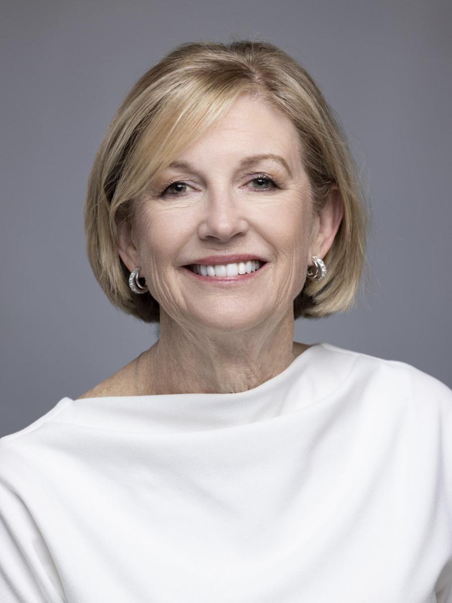 A woman with a classy blonde bob smiles at the camera. She is wearing a solid white blouse with a high neckline; small gold hoop earrings; and professional, natural-looking makeup.