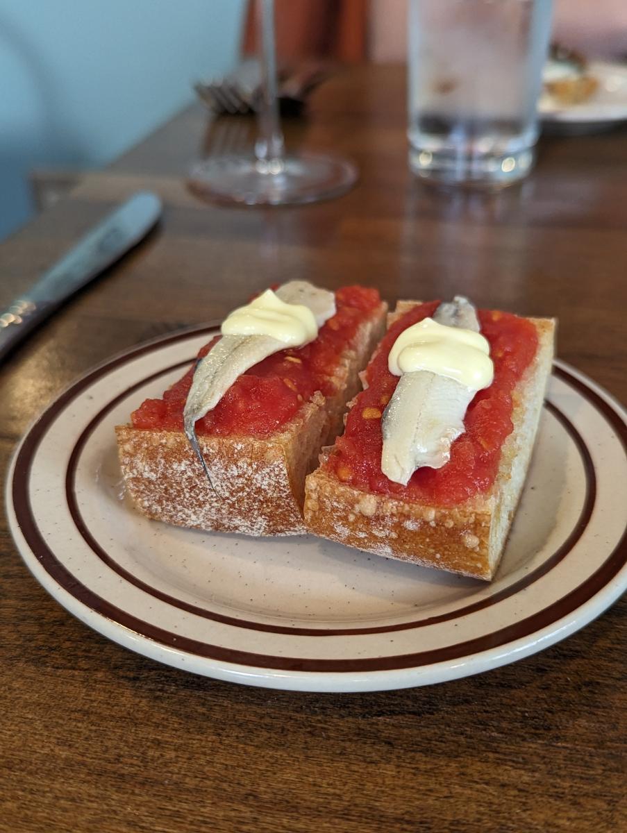 Image is of two pieces of baguette bread with tomato, aioli, garlic and anchovies on it.