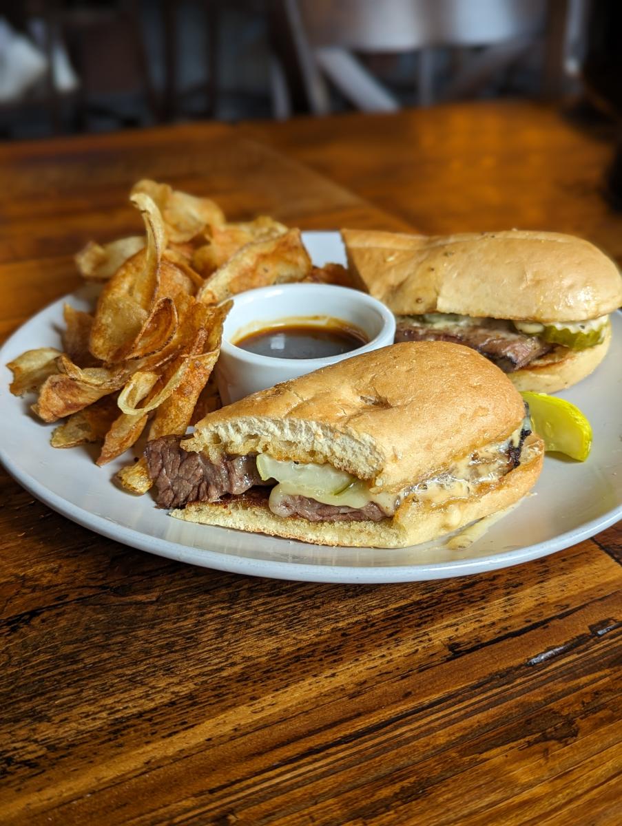 Image is of the brisket sandwich with a pickle, chips and a demi-glace sauce in the center.
