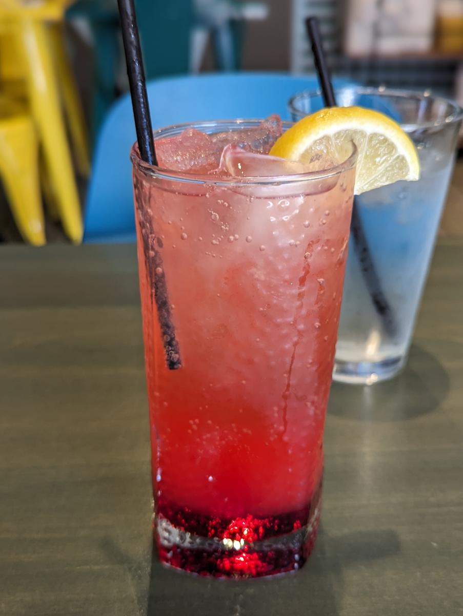 Image is of a red drink in a glass with ice and a lemon wedge.