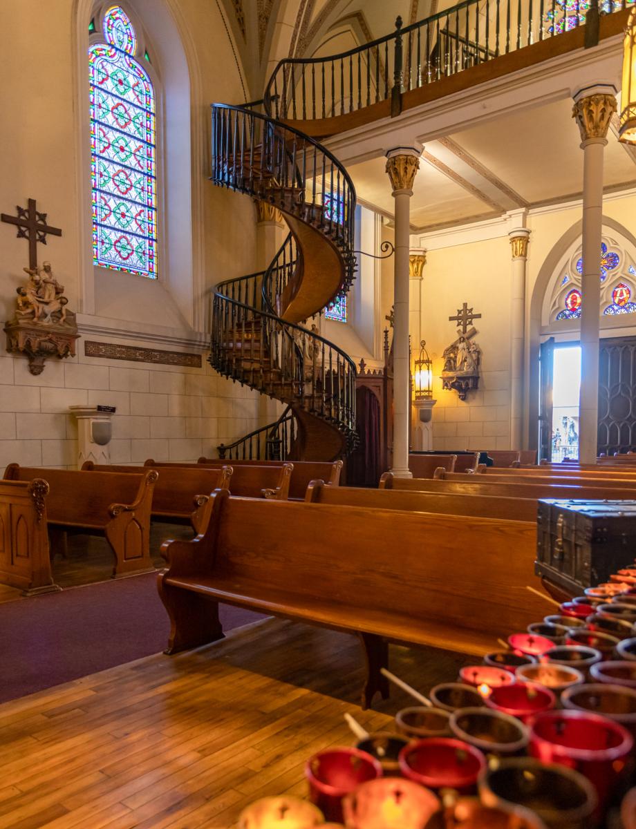 Interior of the Loretto Chapel featuring the famous Miraculous Staircase, with lit votive candles in the foreground and stained glass windows in the background.