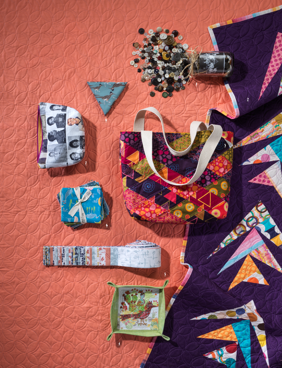 Colorful quilting display on a textured orange backdrop. Includes multicolored quilt, patterned bags, a stack of fabrics, buttons, and a jar.