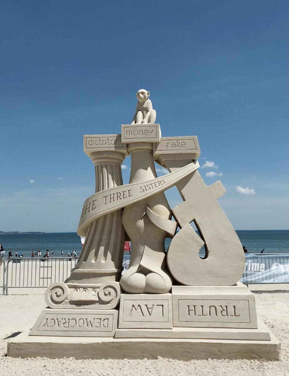 Sand sculpture of three pillars. The bottom says "Truth, Law, and Democracy" upside down. On top, the words say, "Dictator, money, and fake" with a monkey.