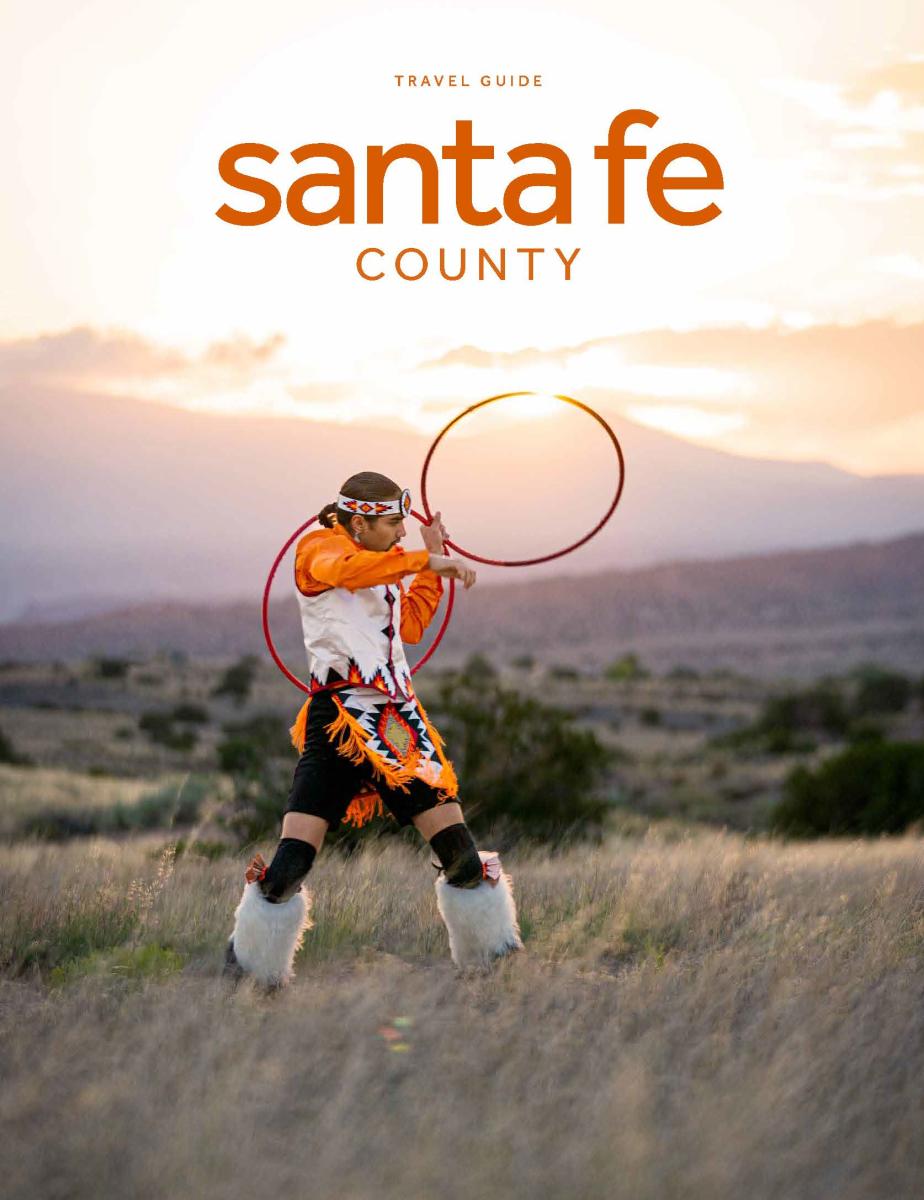 Cover of a Santa Fe County travel guide featuring a person in traditional Native American attire performing a hoop dance at sunset in a scenic desert landscape.