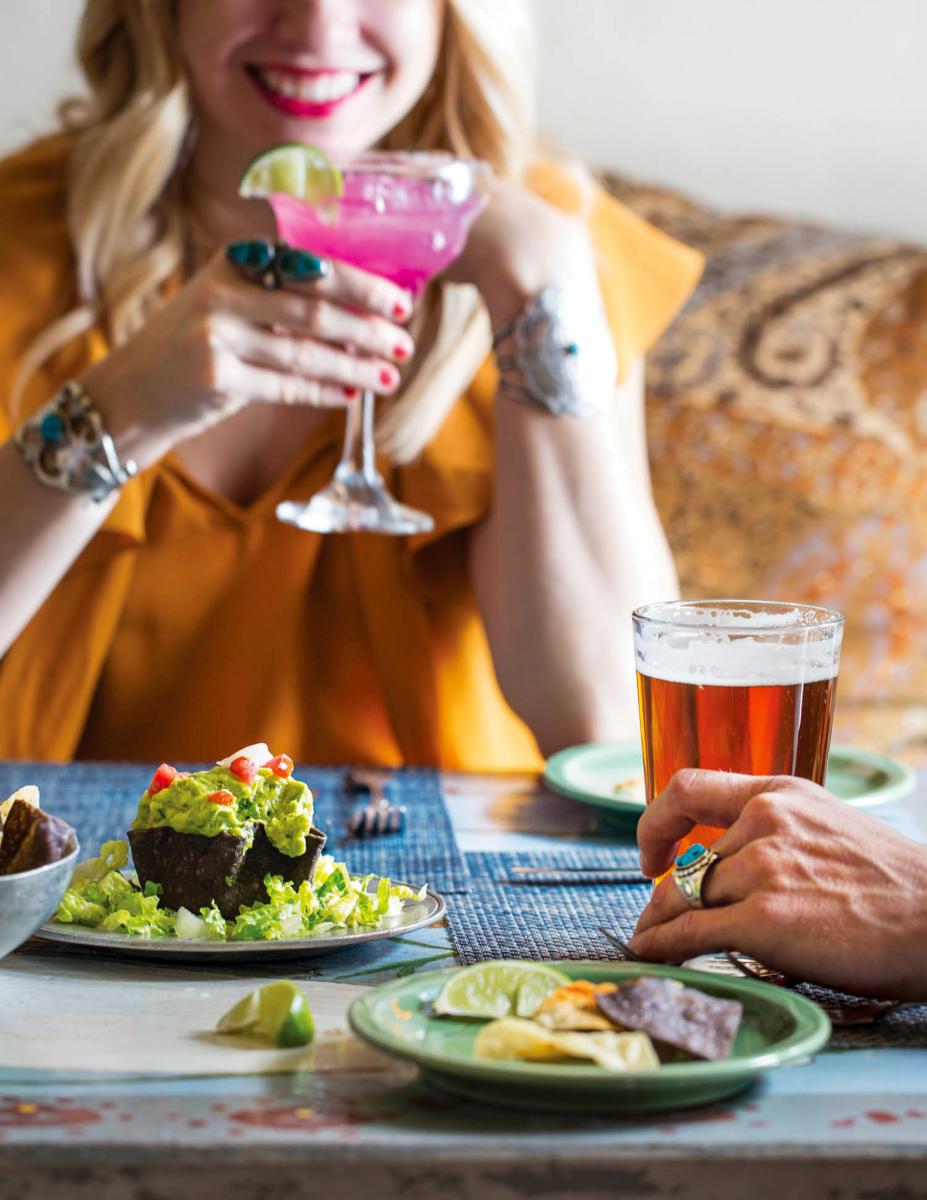 Person holding a pink cocktail at a table with another individual, who has a beer and Mexican food like tacos and guacamole, in a casual dining setting.