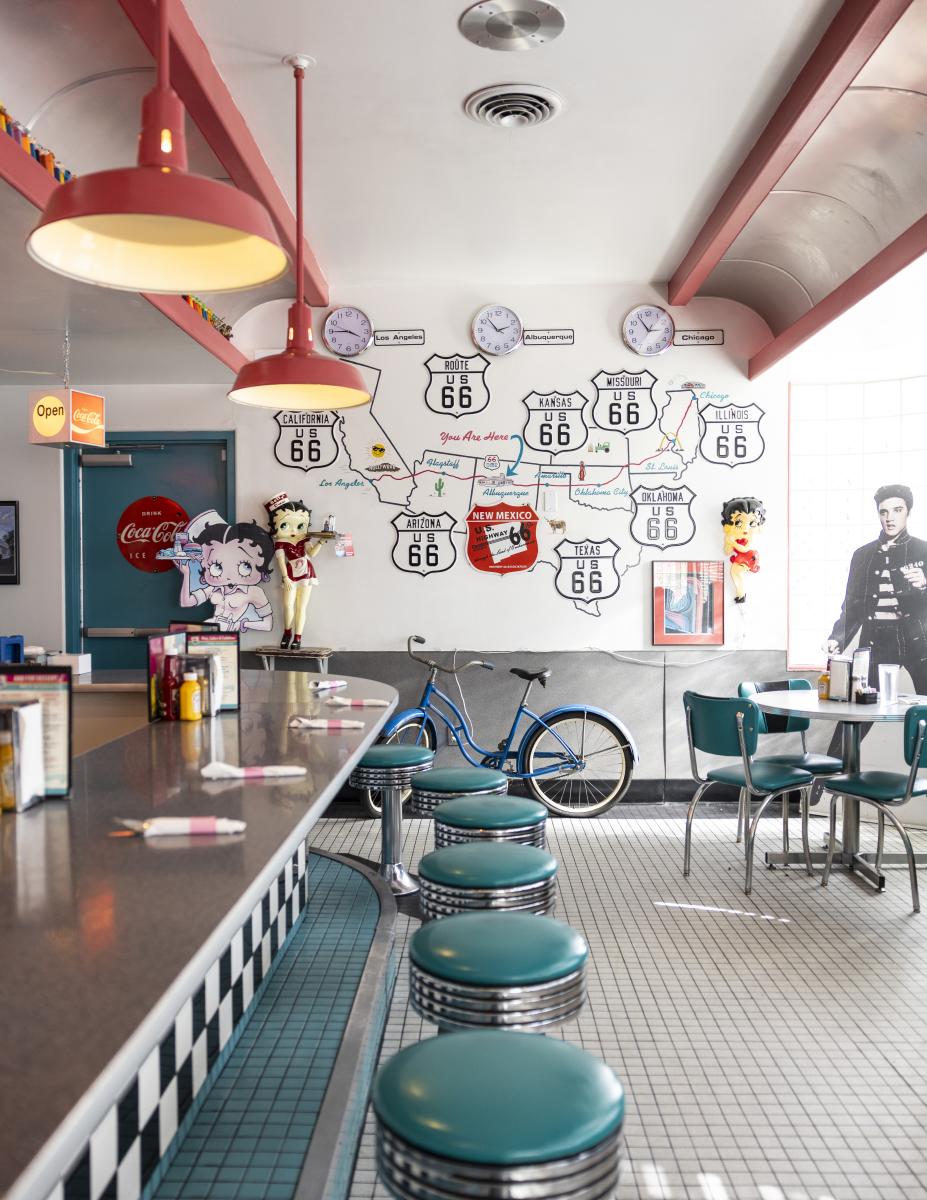 Interior of a retro-themed diner featuring Route 66 and Coca-Cola decorations, teal booths, and a vintage bicycle displayed against a colorful mural wall.