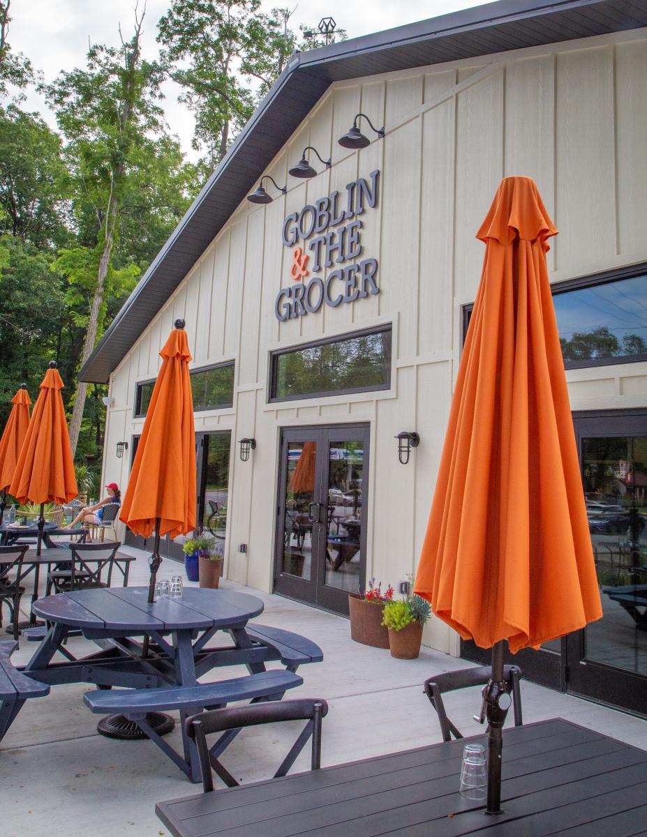 The exterior of "Goblin & The Grocer," a cream-colored building with black trim and a rustic design. The outdoor seating area features dark wooden picnic tables with bright orange umbrellas. Lush greenery and tall trees surround the restaurant, and a few patrons are visible in the background.