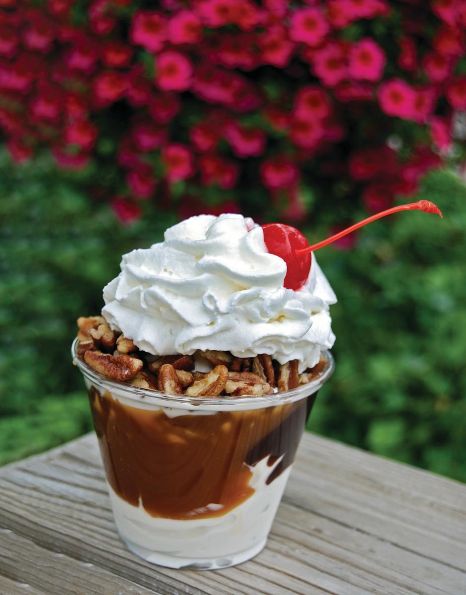 A turtle sundae sits on a wooden table, layered with vanilla frozen custard, hot fudge, caramel, candied pecans, whipped cream and a maraschino cherry. Vibrant pink flowers create a softly blurred backdrop behind the dessert.