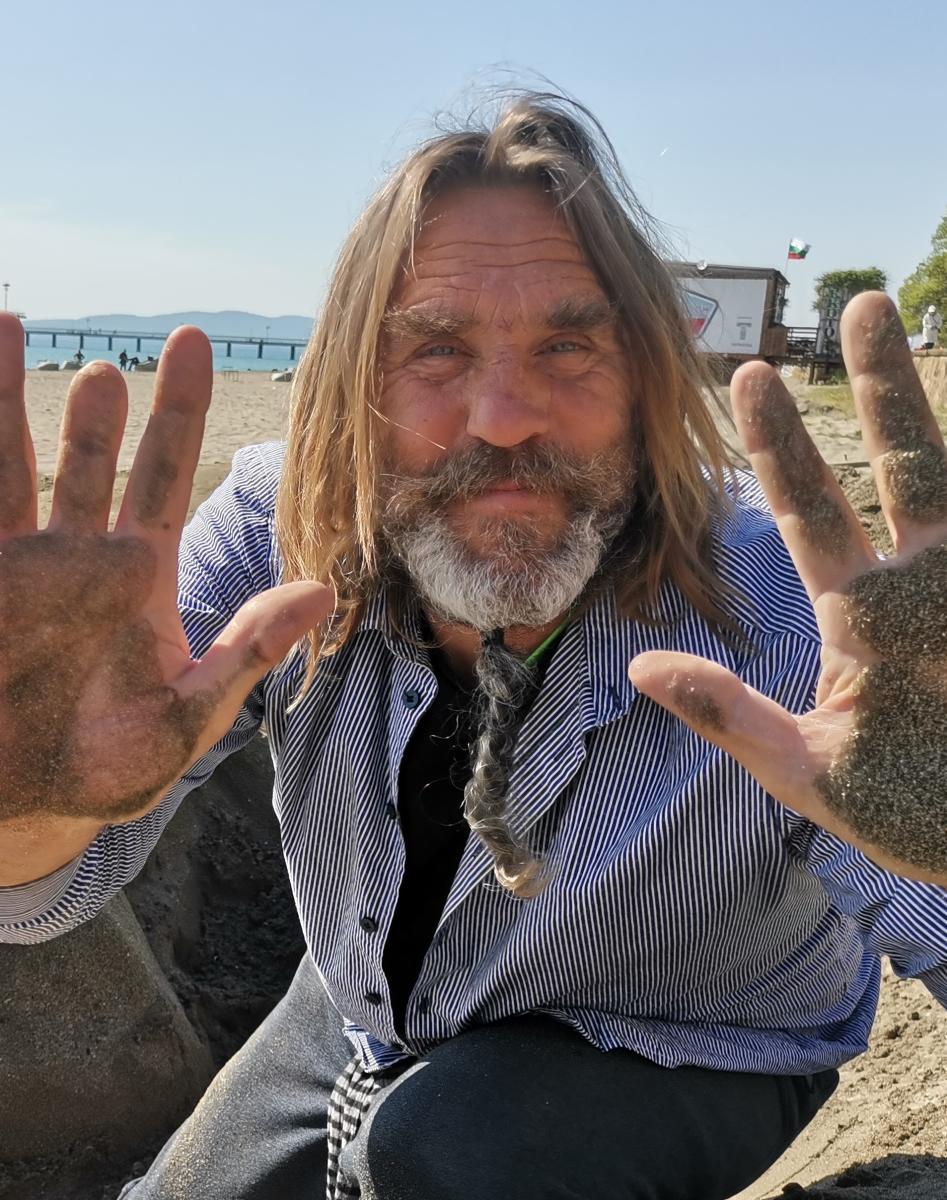 Close up of a man leaning towards the camera, hands spread wide and covered in sand. He has a beard and long hair and is wearing a loose button up while he kneels in the sand.