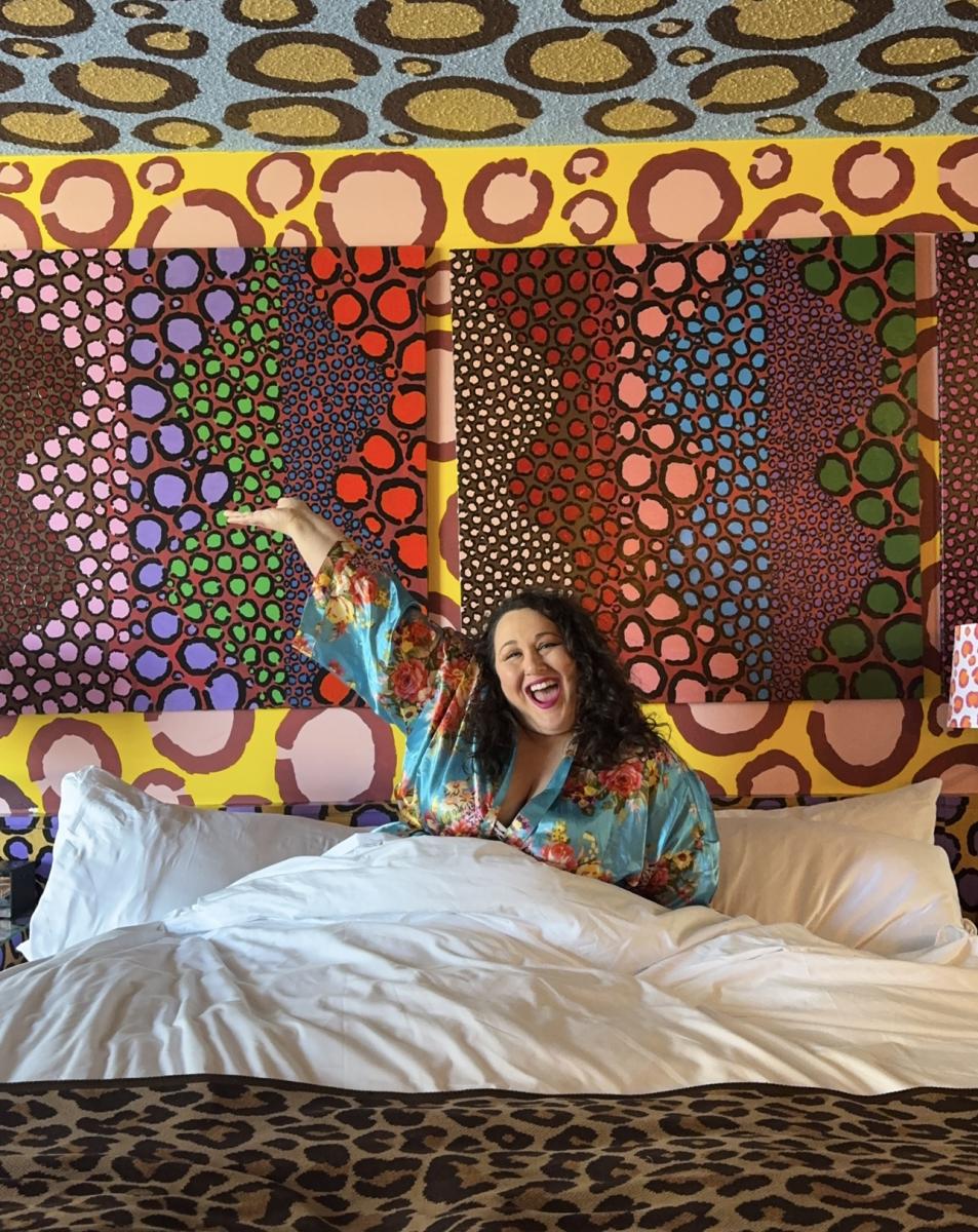 Alysse Delasandro smiles from bed beneath colorful leopard-print artwork at Saint Kate – The Arts Hotel in Milwaukee.