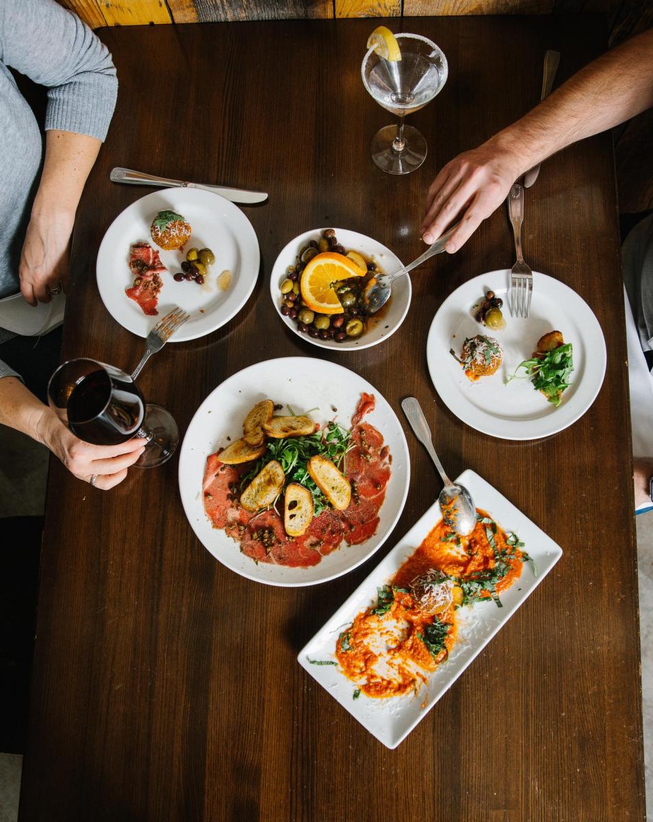 A wooden table displays an array of gourmet dishes, including seafood, salads, and appetizers, with drinks in elegant glasses.