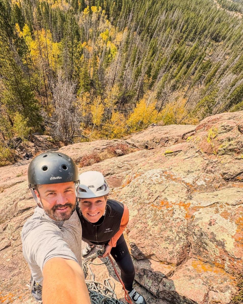 Two climbers smiling at the top of a climbing route in Vedauwoo, Wyoming, with colorful autumn trees and a forested landscape in the background.