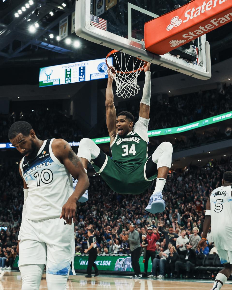 A Milwaukee Bucks player hangs from the rim after a slam dunk as Minnesota Timberwolves defenders walk away, with fans cheering in the background at Fiserv Forum.