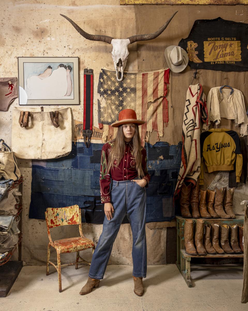 A person in a red cowboy hat and vintage Western attire stands in a rustic room. Decor includes a skull wall mount, vintage flags, and Western boots.