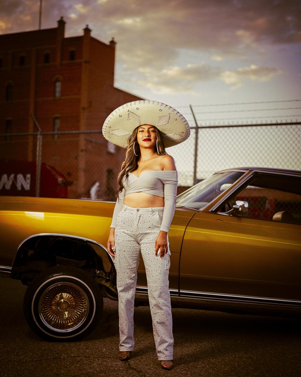 A portrait of Unique Lovato sitting inside her father’s Impala, looking toward the photographer, capturing her connection to generations of lowrider culture.