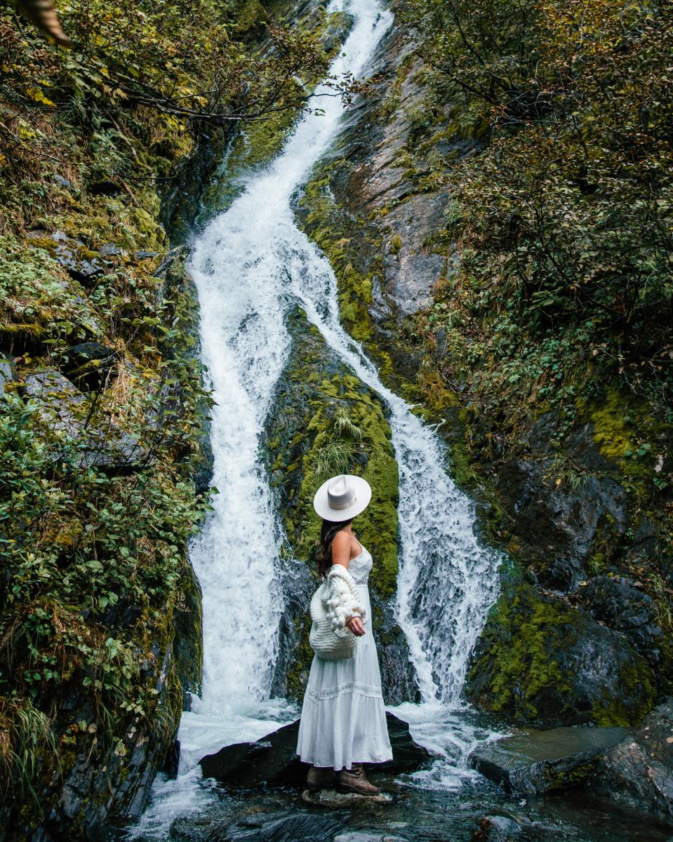 a woman in a brimmed hat and dress standing in front of a waterfall