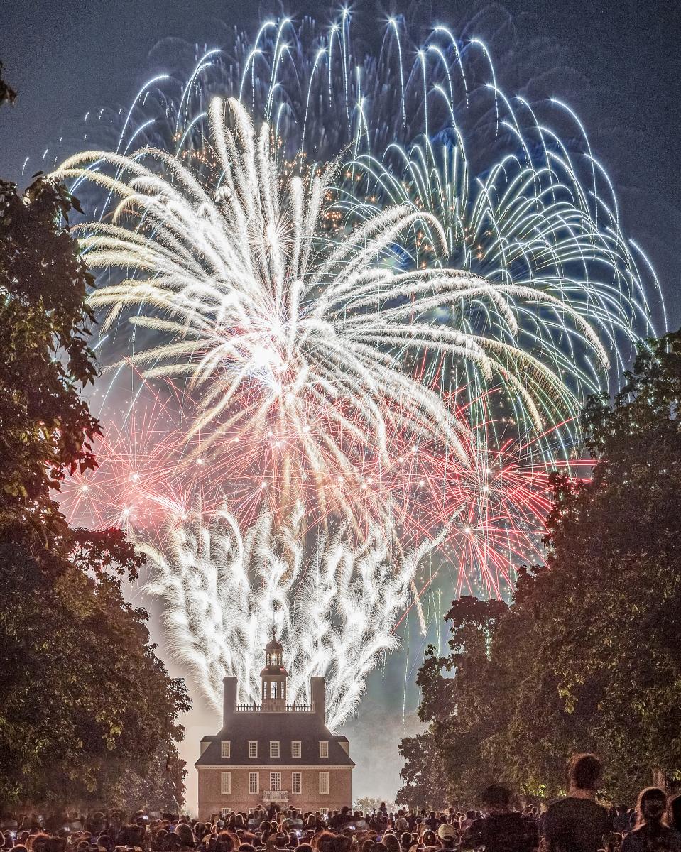 Fireworks illuminate the night sky above a historic building, surrounded by a crowd of spectators. Lush trees frame the scene, enhancing the festive atmosphere.
