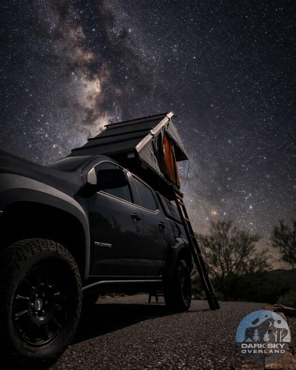 a night photo by @darkskyoverland showing a camper van in the foreground with a tent on top and the desert sky filled with stars and the Milky Way galaxy