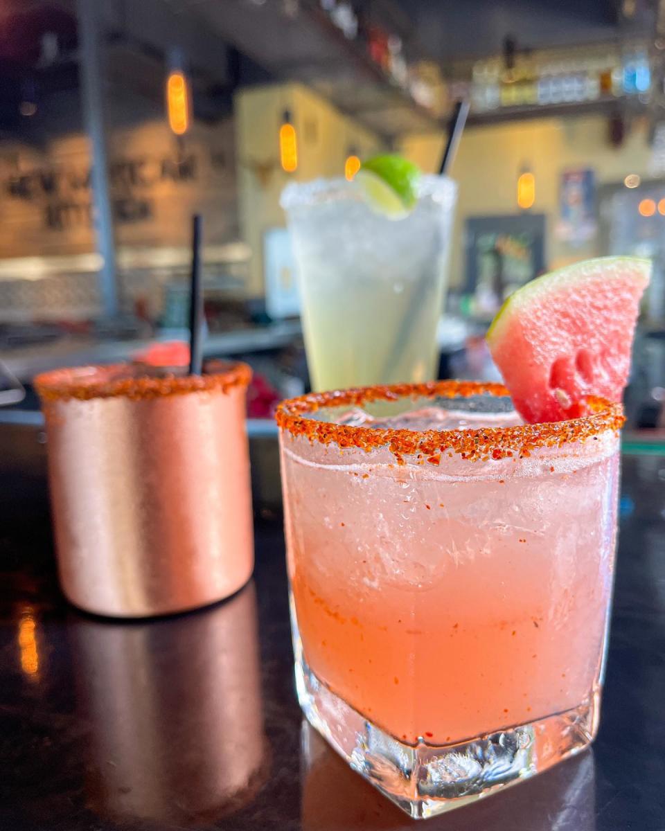 A pink margarita in a short square glass with a tajin rim and watermelon slice for garnish. Two cocktails in a copper mug and tall thin glass can be seen sitting blurred in the background.