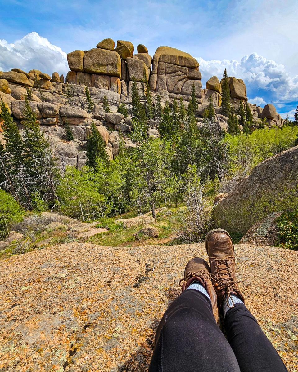 Person relaxing on a rocky ledge with boots visible, overlooking Vedauwoo rock formations and a forested landscape in Wyoming under a blue sky.