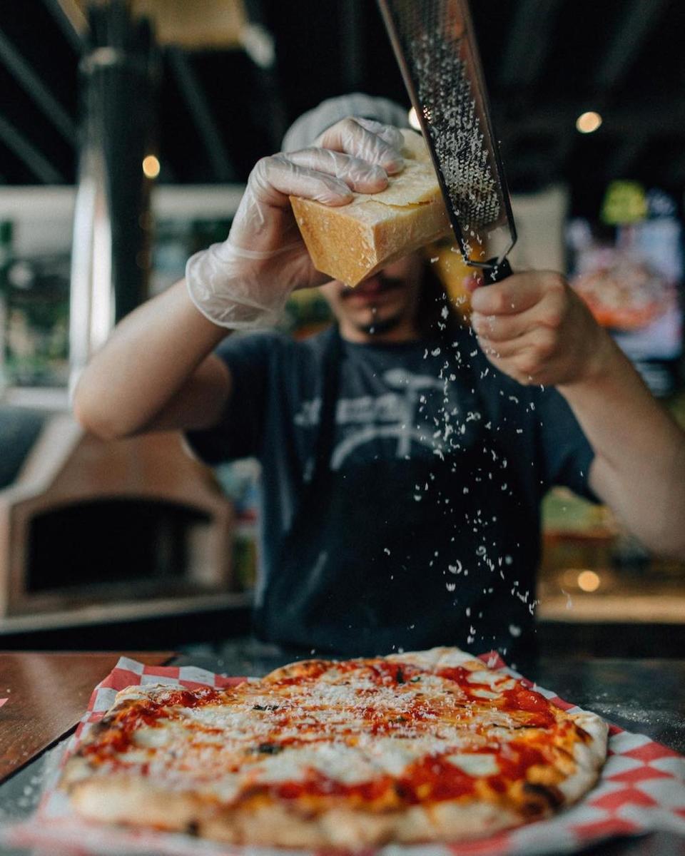 Image is of a guy shaving parmesan cheese onto a woodfired pizza.