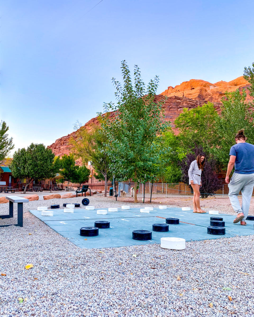Guests playing giant checkers at the shaded grounds of an RV resort with towering red rock cliffs in the background.