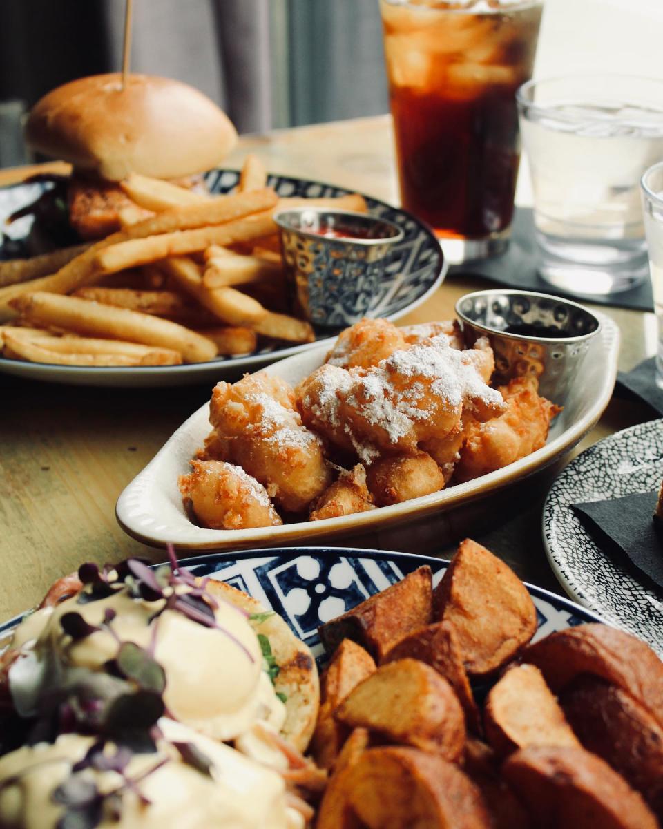 A cozy brunch spread featuring crispy potatoes, eggs Benedict, powdered sugar–dusted fritters with dipping sauce, and a sandwich with fries, all paired with iced tea and water on a sunlit table.