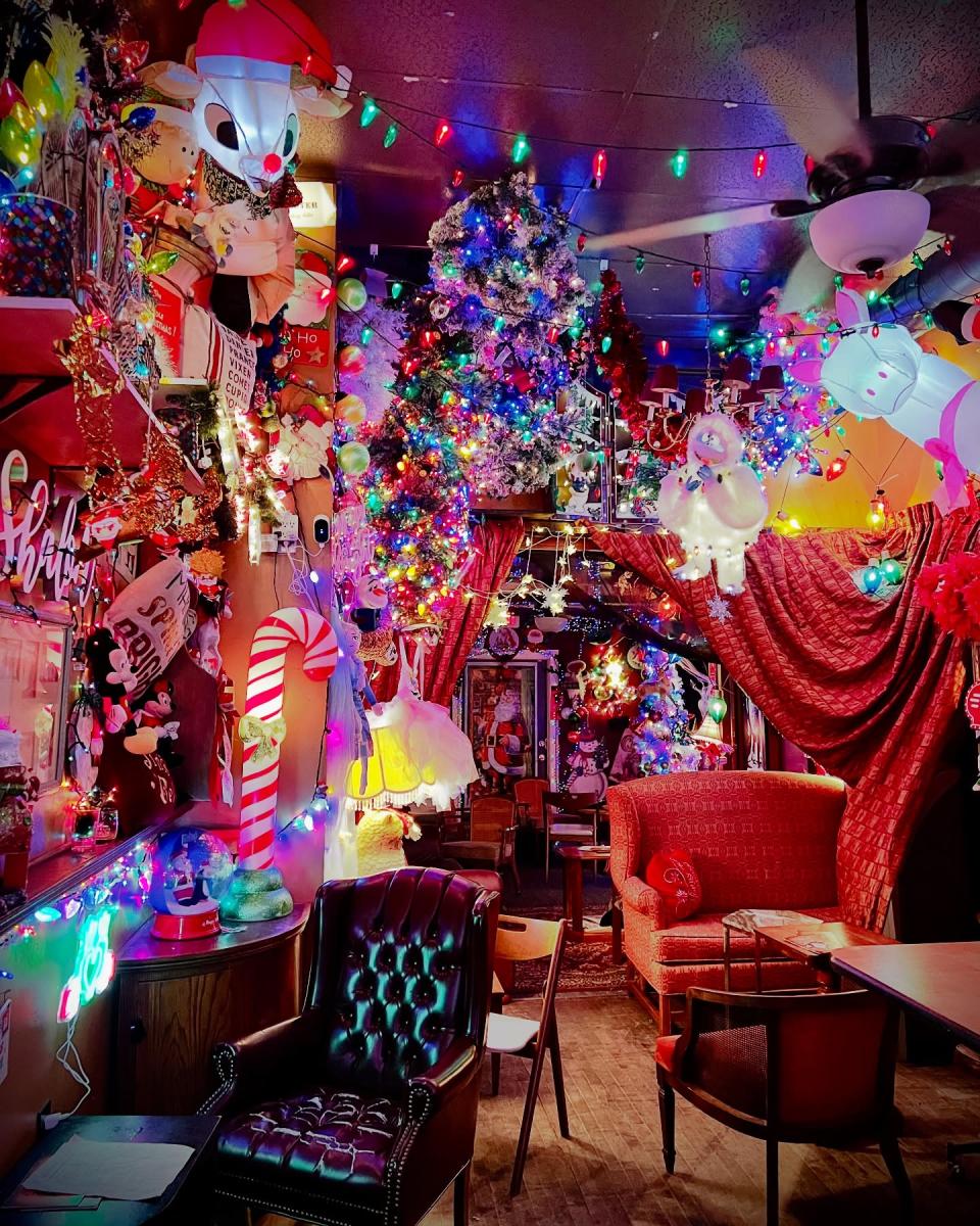 A room filled with dense holiday decorations including glowing string lights, ornaments, garlands, and inflatable characters that cover the walls and ceiling. A brightly lit Christmas tree hangs at an angle near the center of the space. Plush figures, candy cane props, and colorful light displays create a playful and crowded scene. Vintage style chairs and small tables sit beneath heavy red curtains. The entire room feels packed with holiday items that surround the seating area with color and light.