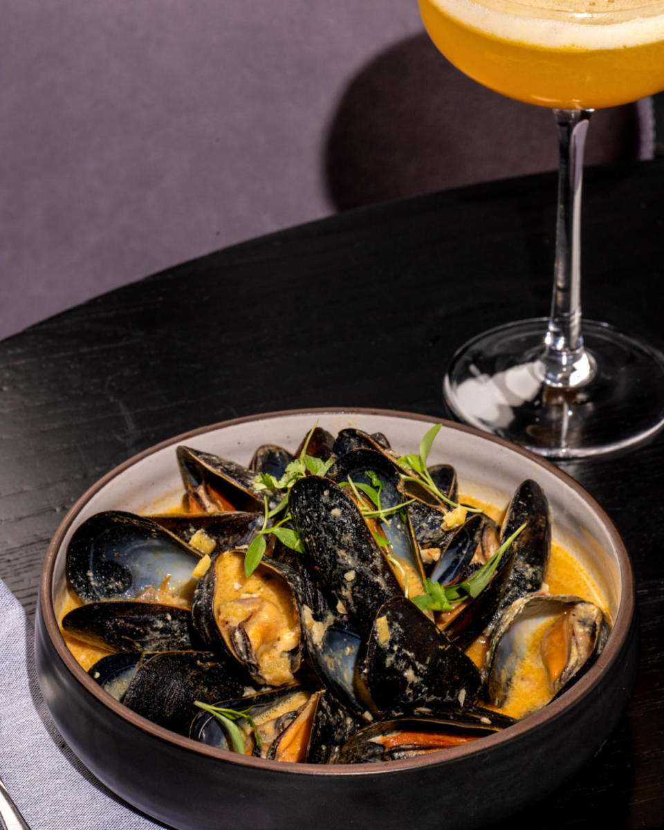 Bowl of mussels in a creamy orange broth garnished with fresh herbs at The Conroy, with a golden cocktail served alongside.